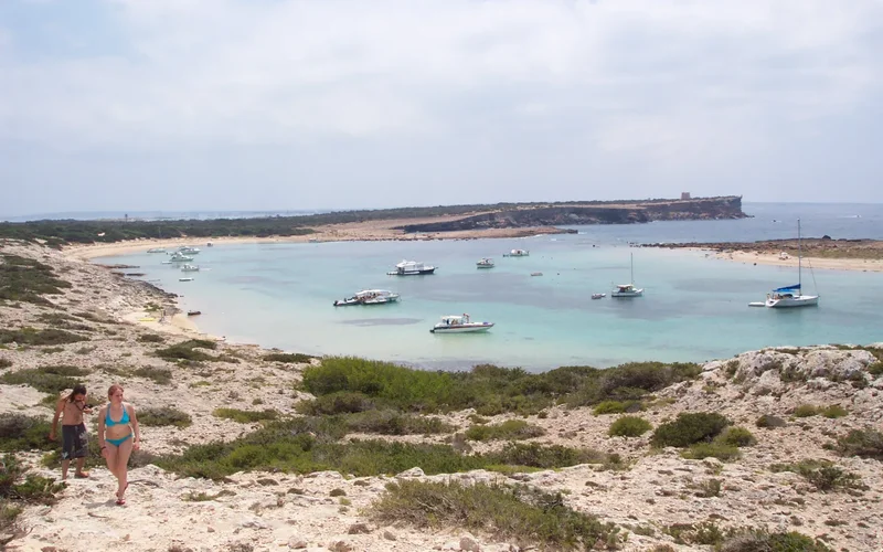 Playa de arena blanca en Formentera con aguas turquesas, barcos anclados y una pareja caminando por la orilla rocosa bajo un cielo nublado