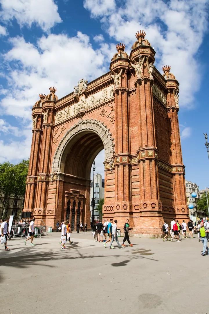 El Arco del Triunfo de Barcelona, de ladrillo rojo ornamentado, bajo un cielo azul con nubes, con personas caminando en la plaza