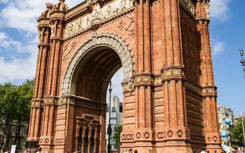 El Arco del Triunfo de Barcelona, de ladrillo rojo ornamentado, bajo un cielo azul con nubes, con personas caminando en la plaza