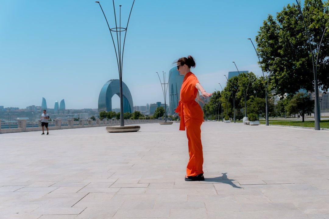 Mujer con mono naranja y gafas de sol posa con brazos extendidos en el boulevard de Bakú, ante torres Flame, arco moderno y mar Caspio bajo cielo azul