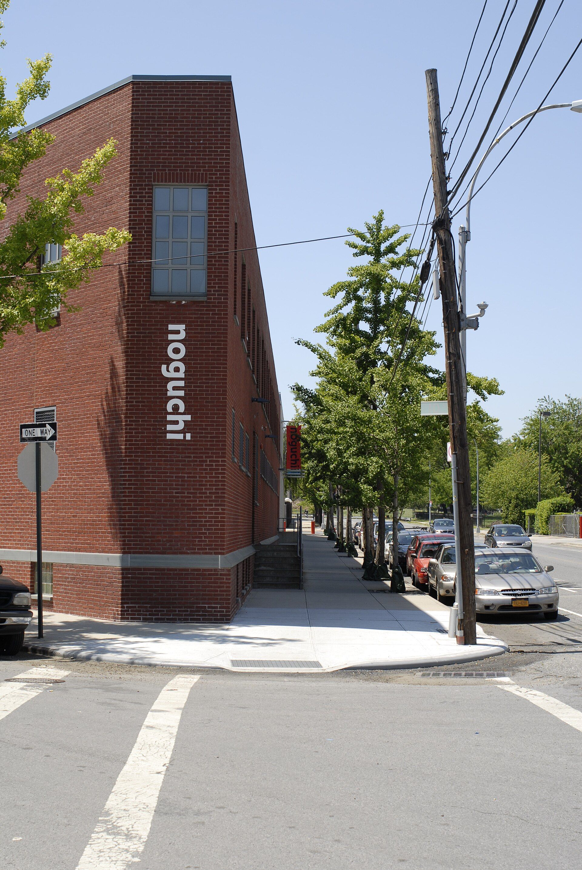 Edificio de ladrillo rojo con el nombre 'noguchi' en vertical, junto a una acera con árboles y coches aparcados.