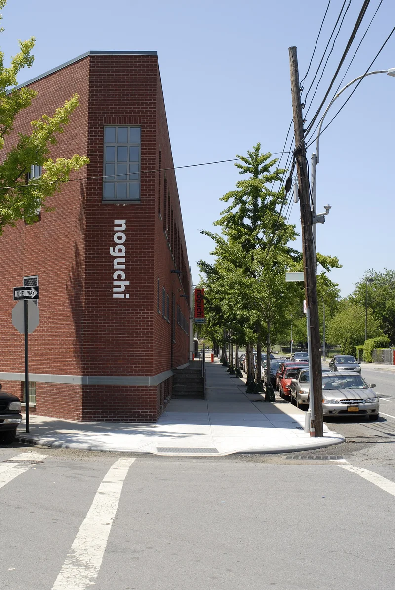 Edificio de ladrillo rojo con el nombre 'noguchi' en vertical, junto a una acera con árboles y coches aparcados.