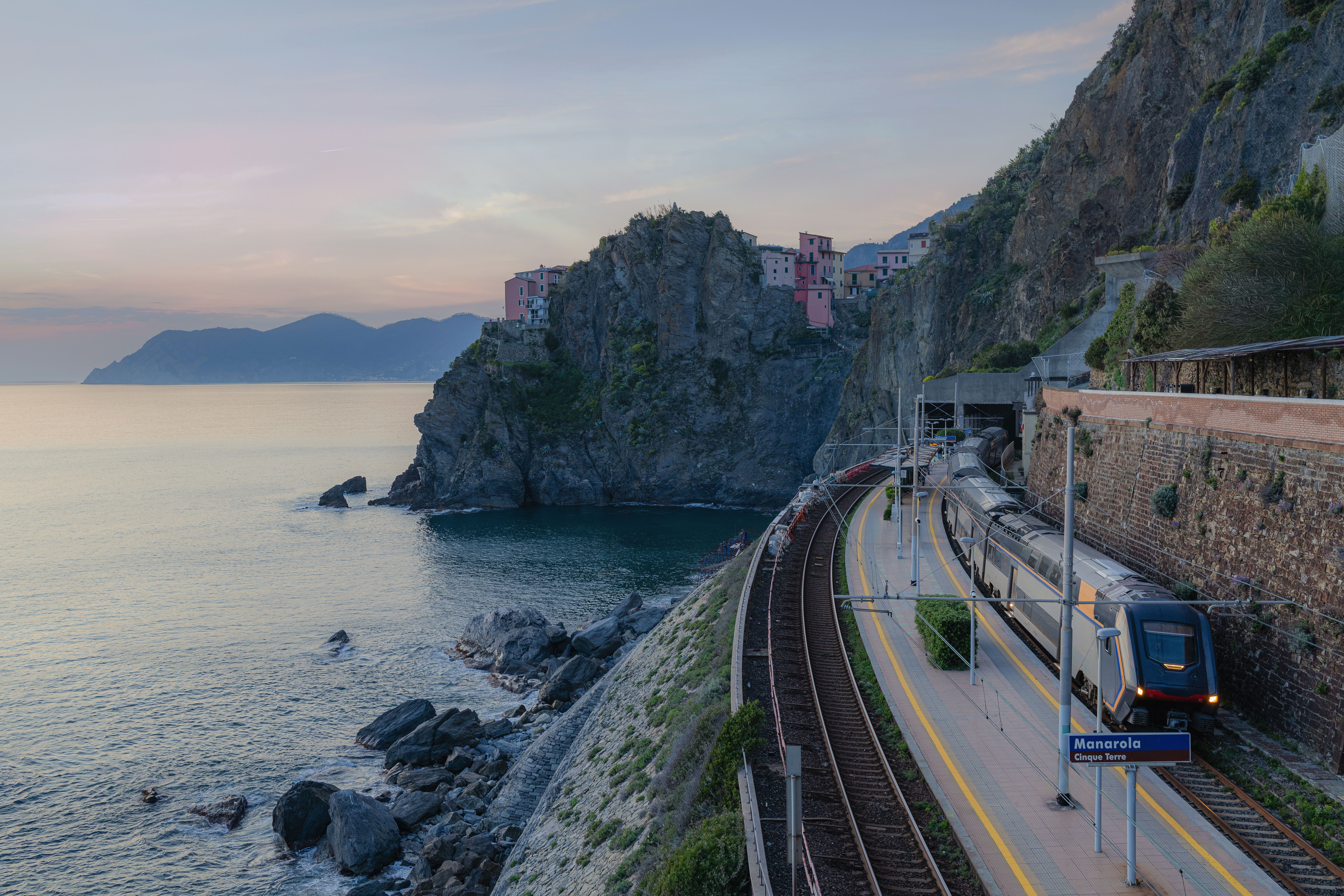 Tren en una estación costera con un pueblo en un acantilado y el mar al atardecer.