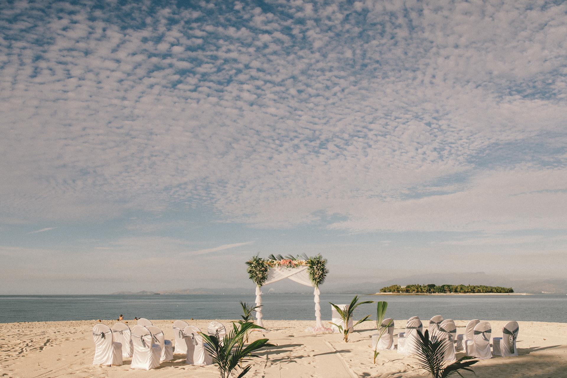 Escena de boda en playa de arena blanca con arco floral blanco, sillas cubiertas alineadas, palmeras y mar azul bajo cielo nublado