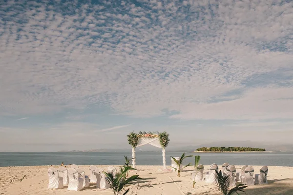 Escena de boda en la playa de Nadi, Fiyi, con arco floral blanco, sillas cubiertas, palmeras y vista al mar con isla bajo cielo nublado