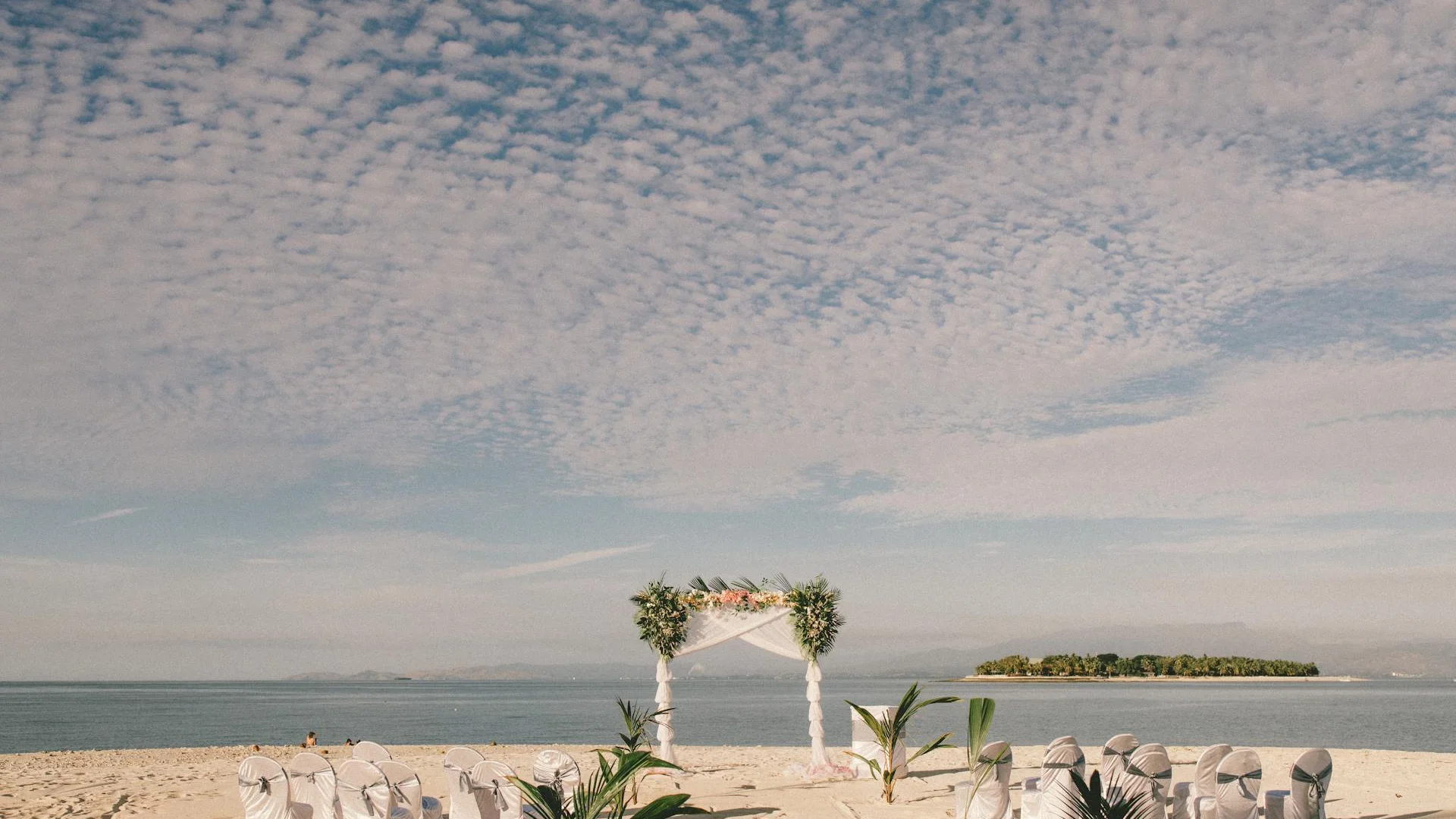 Escena de boda en la playa de Nadi, Fiyi, con arco floral blanco, sillas cubiertas, palmeras y vista al mar con isla bajo cielo nublado