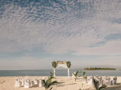 Escena de boda en playa de arena blanca con arco floral blanco, sillas cubiertas alineadas, palmeras y mar azul bajo cielo nublado