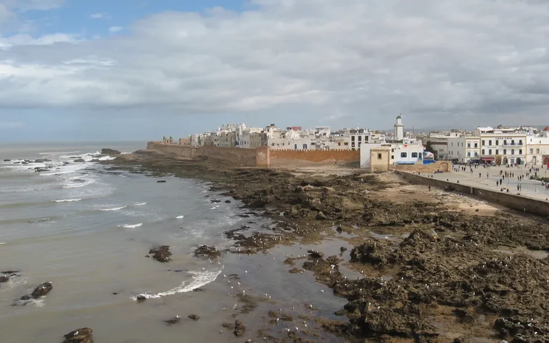Vista de las murallas rojizas de Essaouira junto al Atlántico, con edificios blancos, faro y olas en rocas bajo cielo nublado