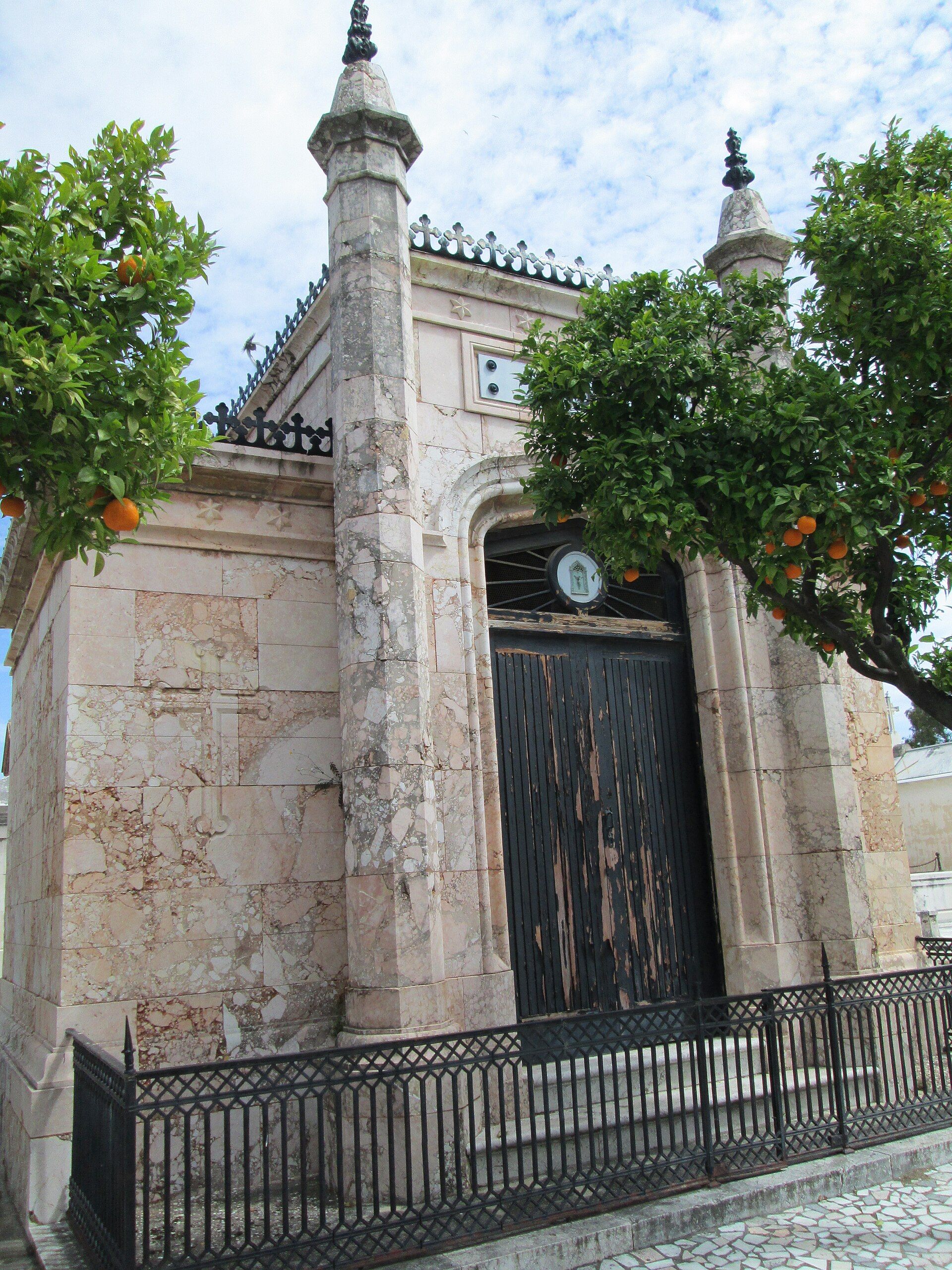 Mausoleo de mármol con puerta de madera oscura, flanqueado por árboles de naranjas bajo cielo nublado.