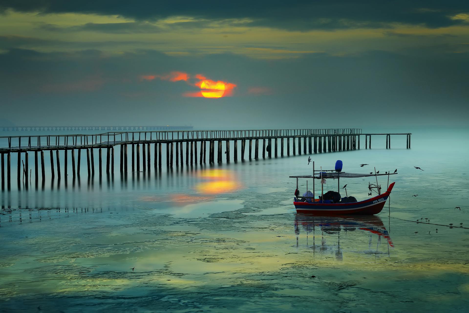 Atardecer sobre un muelle de madera en aguas tranquilas de Penang, con un barco tradicional rojo anclado y reflejos dorados