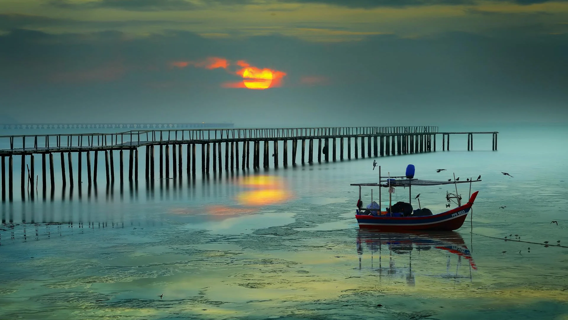 Atardecer en Penang con muelle de madera, barco rojo tradicional y reflejos dorados en el agua brumosa