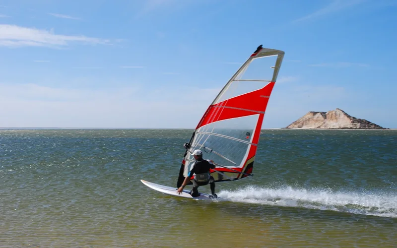 Windsurfista surfeando en el mar turquesa de Dakhla con vela roja y blanca, cielo azul y formación rocosa al fondo