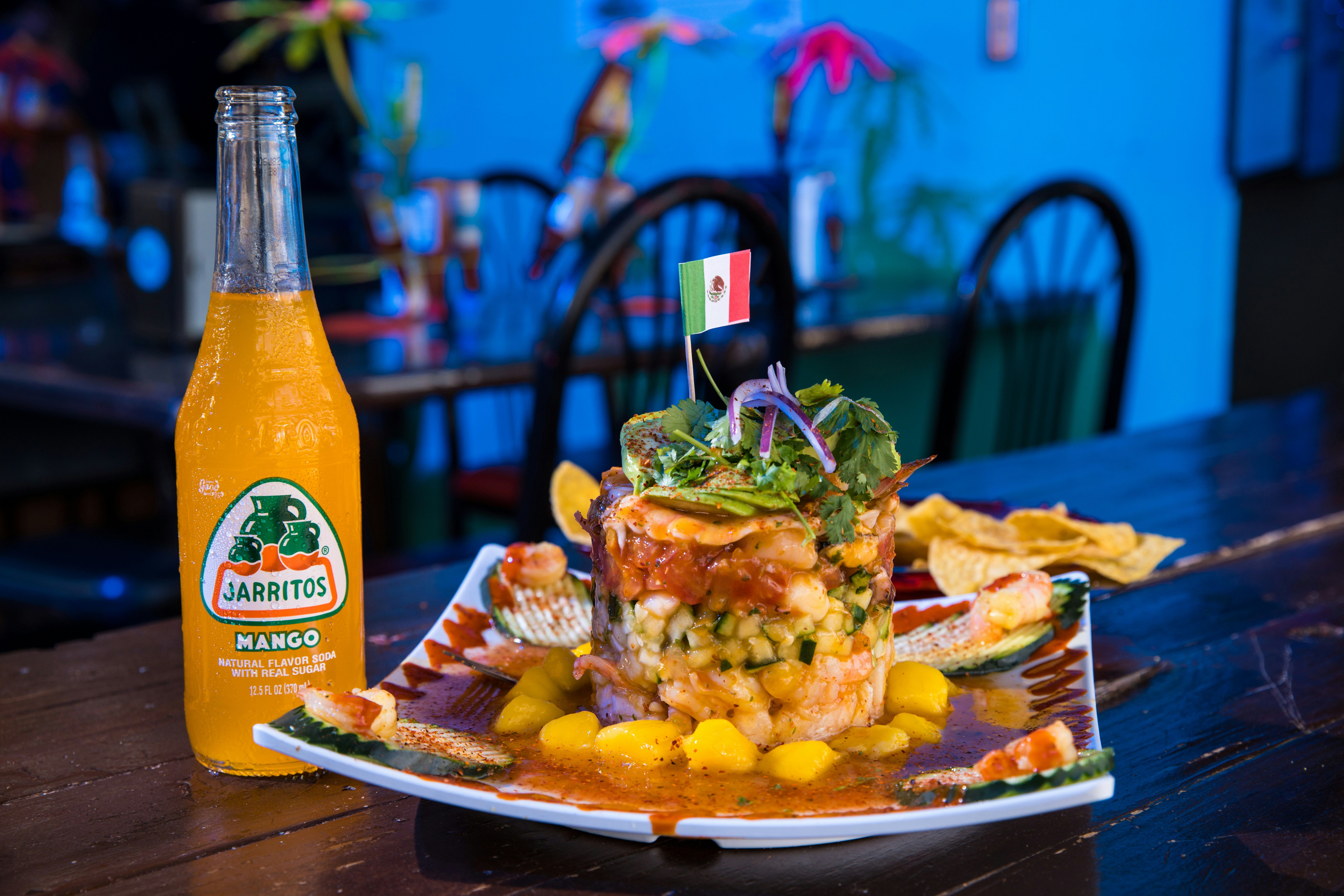Un plato de ceviche con una bandera mexicana, acompañado de una botella de refresco de mango.