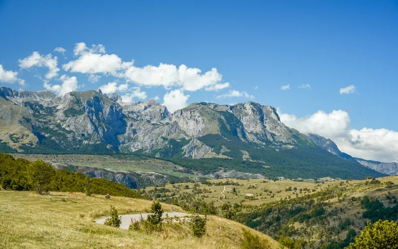 Paisaje montañoso con picos rocosos, valles verdes y un cielo azul con nubes dispersas.