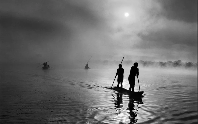 Dos siluetas de hombres con pértigas en una canoa navegan por un río envuelto en niebla, con el sol naciente entre nubes grises