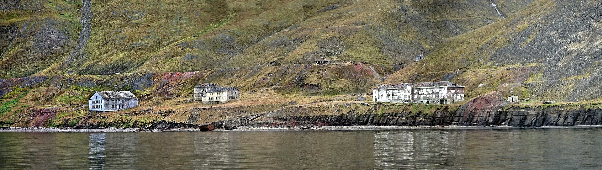 Asentamiento abandonado de Grumant en Svalbard, con edificios ruinosos junto a la orilla y colinas áridas.