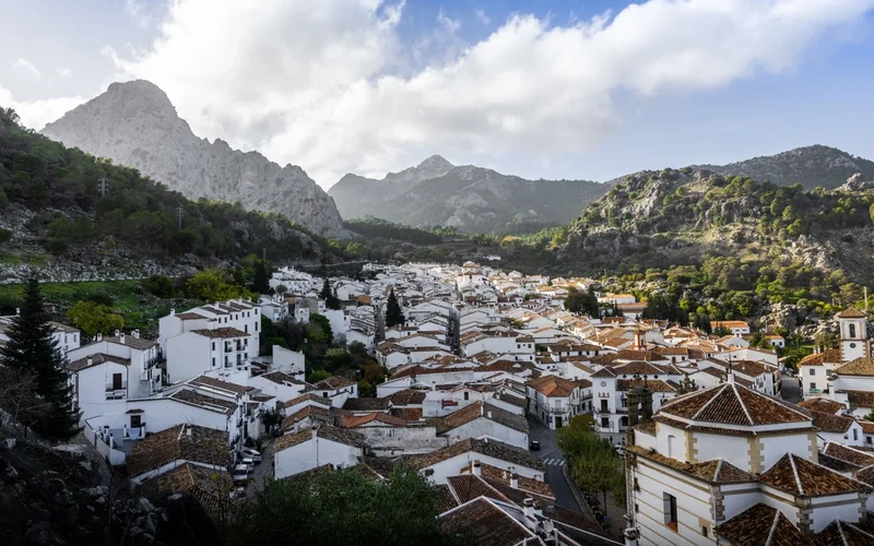 Vista aérea de un pueblo blanco con tejados de terracota, rodeado de montañas verdes y cielo nublado.
