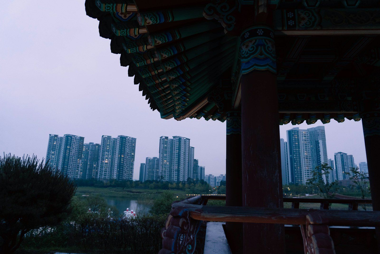 Pabellón tradicional coreano con techo curvado verde y pilares de madera, vista a un río con vegetación y rascacielos modernos en Incheon al atardecer