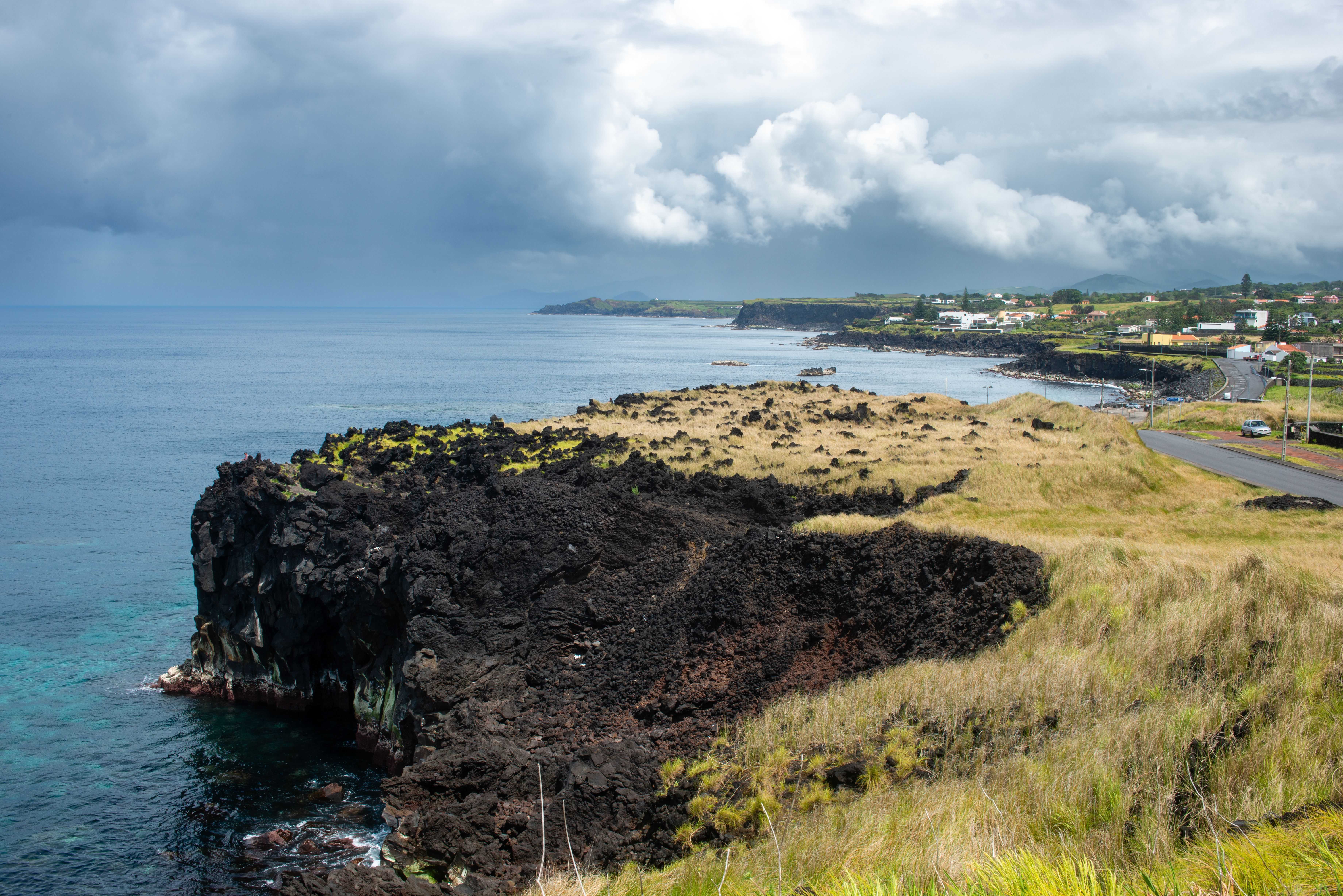 Vista aérea de una costa rocosa con acantilados de lava negra, mar azul y un pueblo a lo lejos.