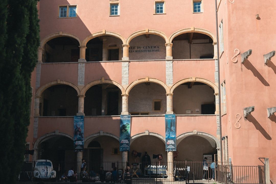 Edificio histórico de fachada rosada con arcos y balcones en un patio soleado, personas paseando junto a un coche blanco y carteles azules colgando