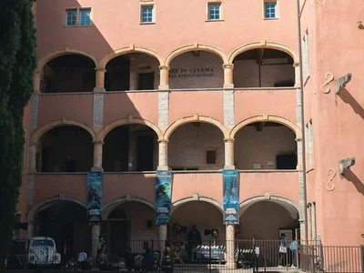 Edificio histórico de fachada rosada con arcos y balcones en un patio soleado, personas paseando junto a un coche blanco y carteles azules colgando