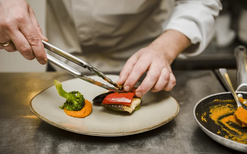 Manos de un chef en mangas blancas usan pinzas para colocar brócoli, pimiento rojo, berenjena y puré naranja en un plato gris sobre encimera metálica