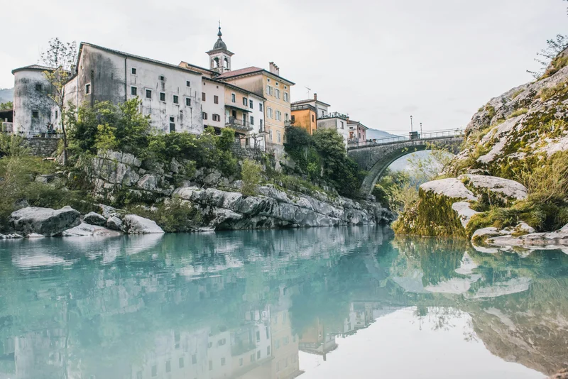 Pueblo con edificios antiguos junto a un río de aguas turquesas y un puente de piedra.