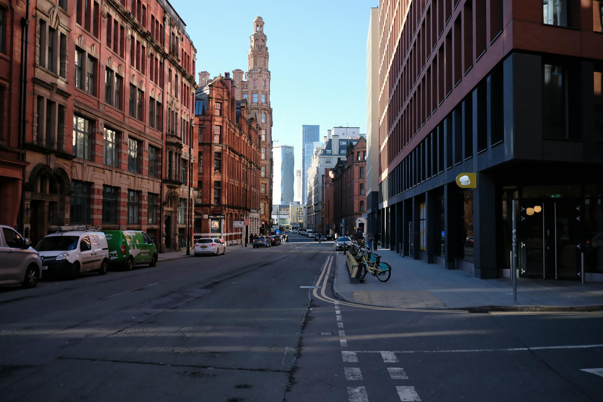 Vista de una calle en Manchester con edificios victorianos de ladrillo rojo a un lado y modernos al otro, autos y bicicletas estacionados bajo cielo
