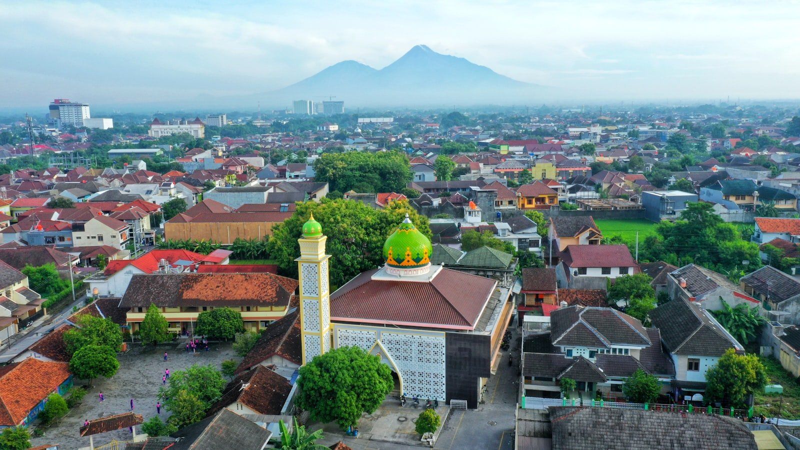 Vista aérea de Yogyakarta con mezquita de cúpula verde, edificios tradicionales rojos y montañas nubladas al fondo