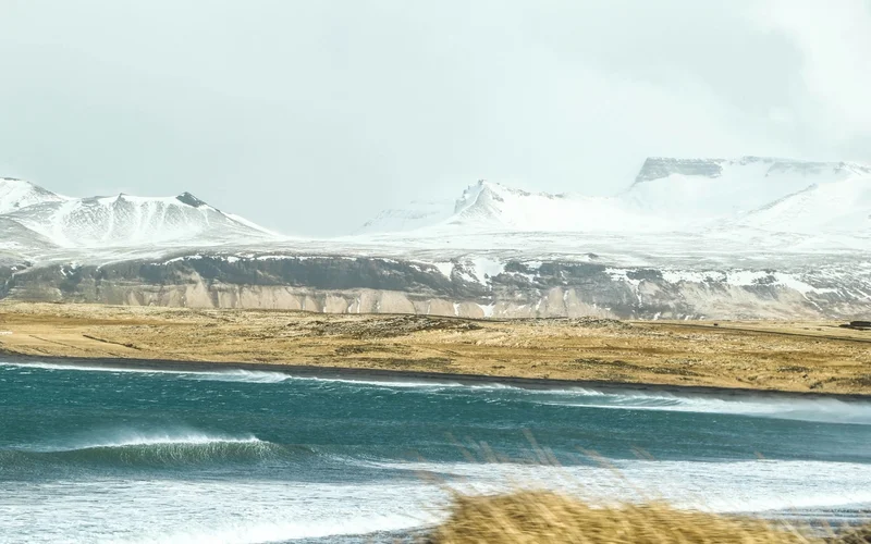 Paisaje costero con montañas nevadas al fondo, playa de arena y mar agitado.