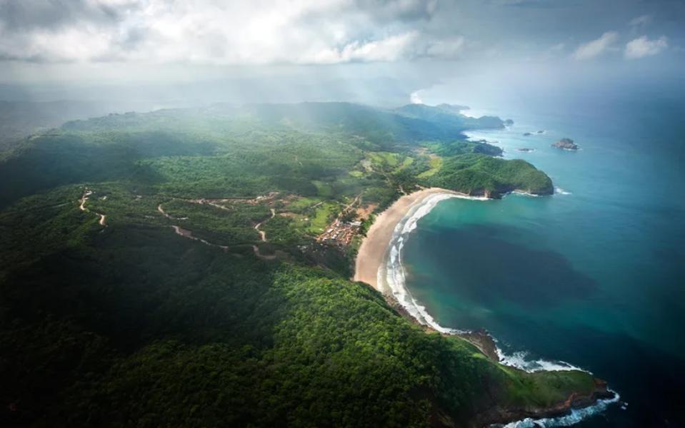 Vista aérea de la playa curvada de Costa Esmeralda, rodeada de selva verde, acantilados y aguas turquesas bajo nubes