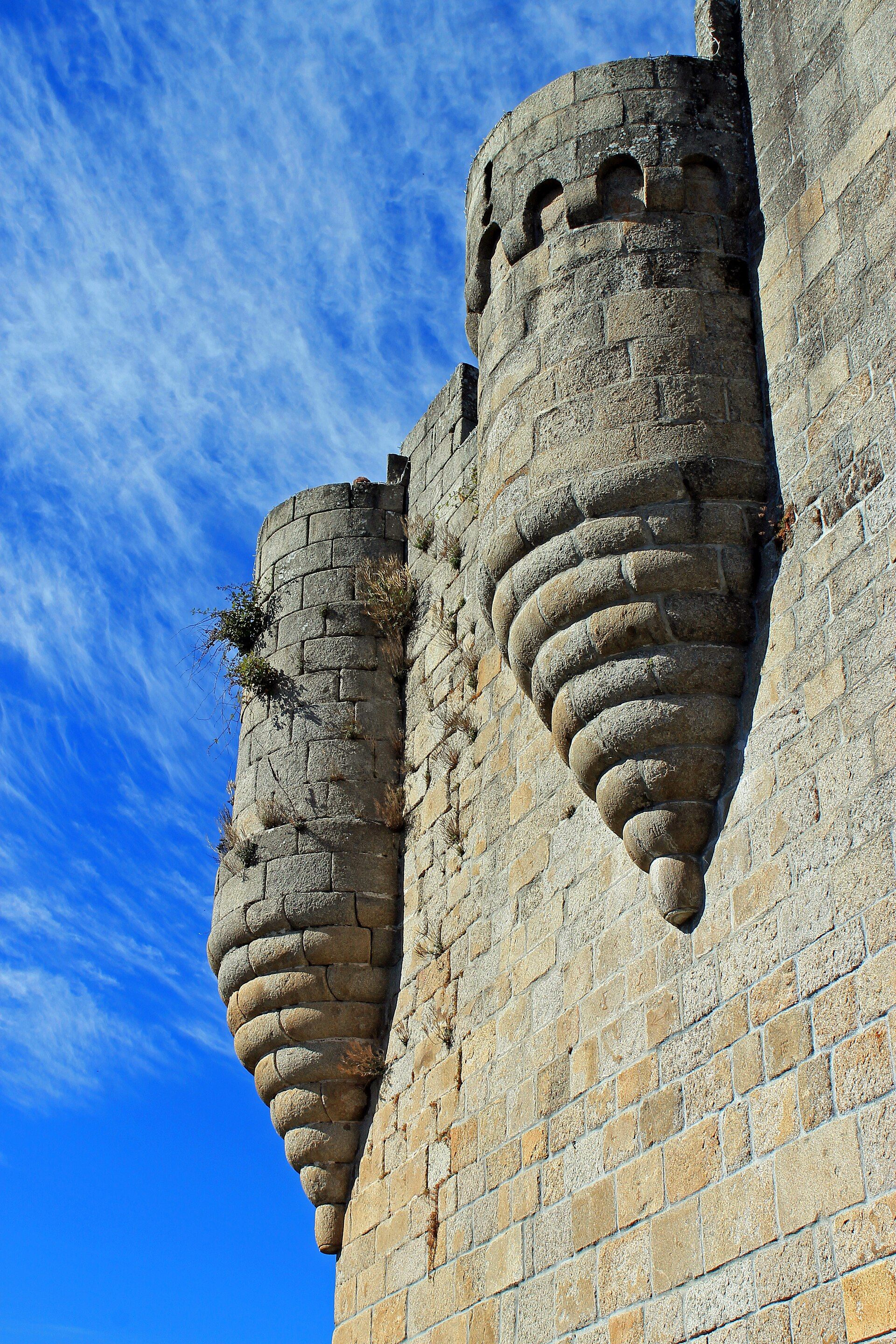 Primer plano de dos torres de piedra de un castillo medieval con almenas y saeteras, bajo un cielo azul con nubes blancas.