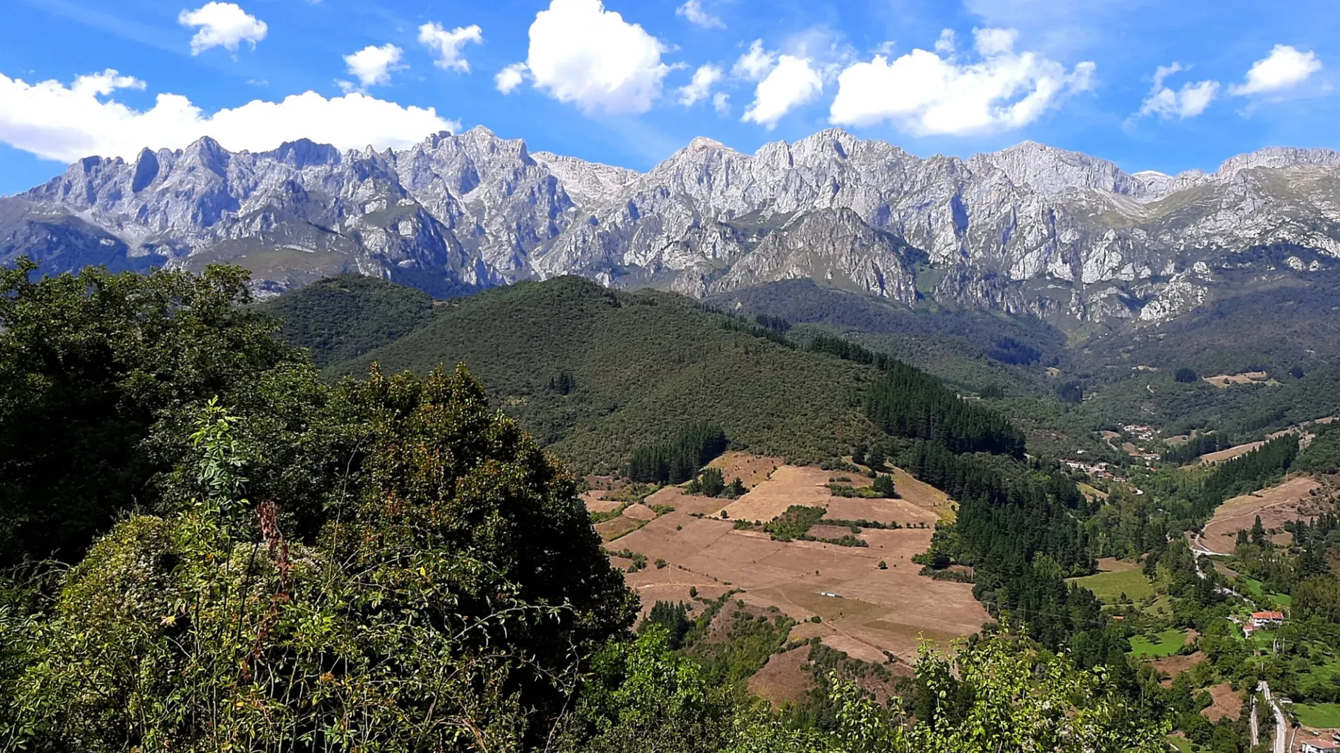 Panorama de los Picos de Europa con picos rocosos nevados, cielo azul nublado, bosques verdes y campos dorados en el valle