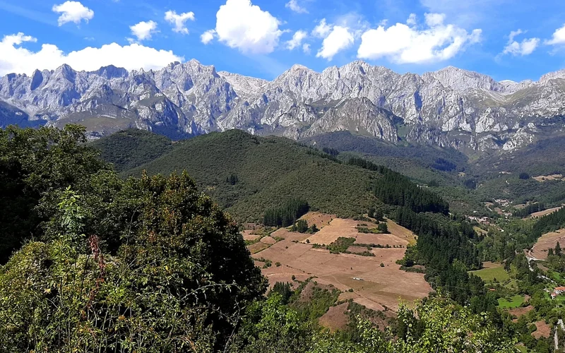 Panorama de los Picos de Europa con picos rocosos nevados, cielo azul nublado, bosques verdes y campos dorados en el valle
