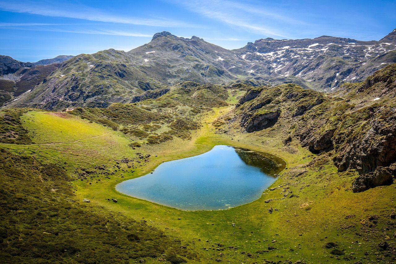 Vista aérea de un lago de montaña rodeado de prados verdes y montañas rocosas con nieve.