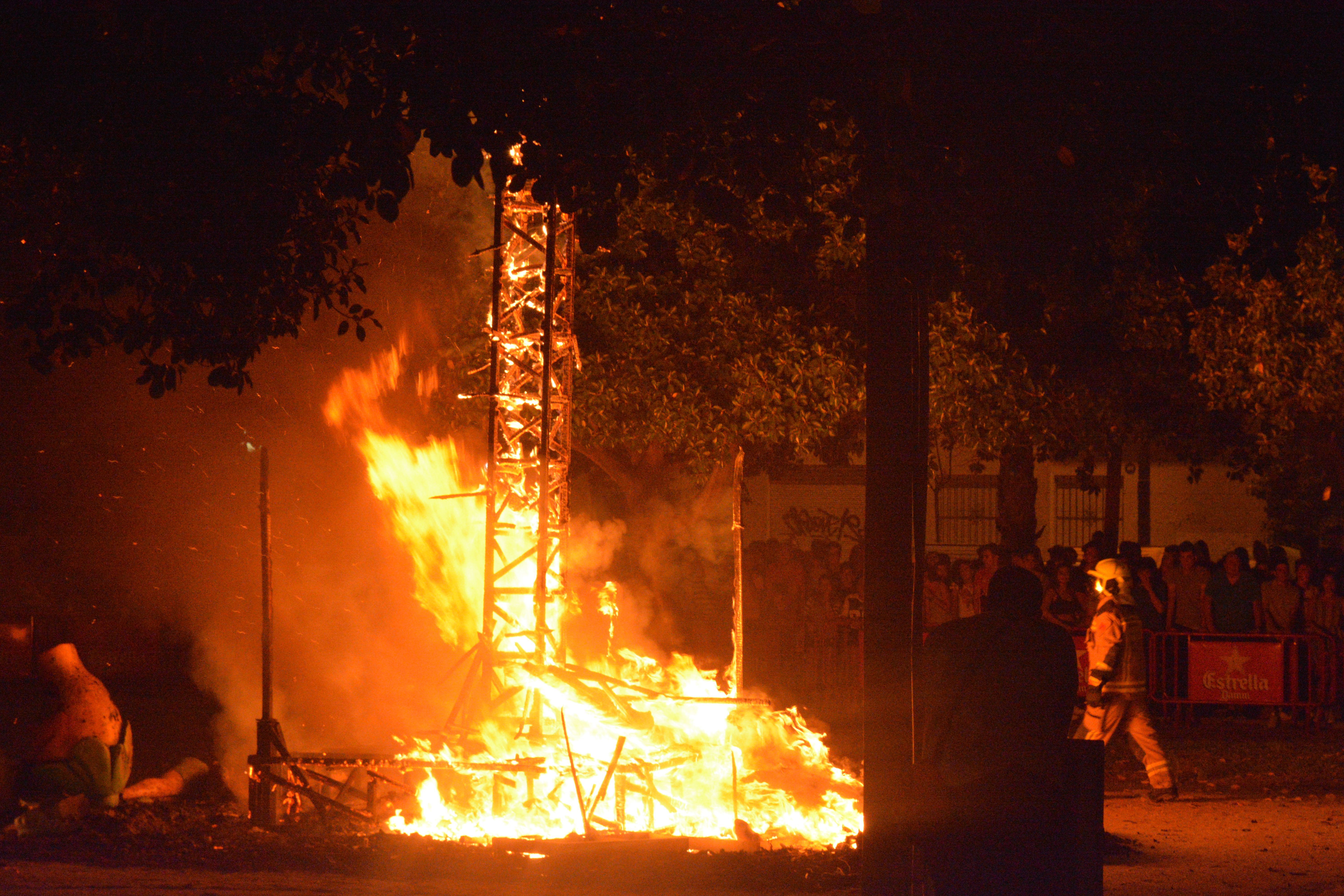 Gran hoguera nocturna con llamas intensas y una estructura de madera ardiendo, con personas observando.
