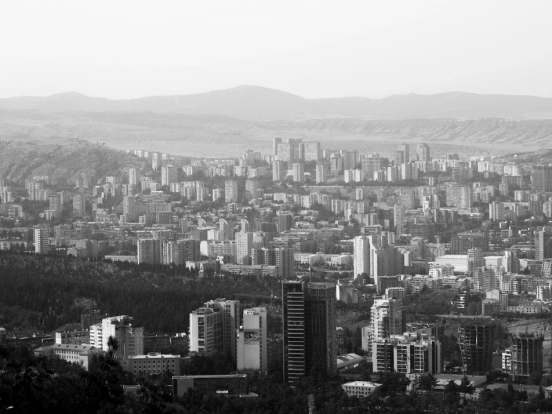 Vista panorámica en blanco y negro de Tiflis, con rascacielos densos en colinas y montañas al fondo