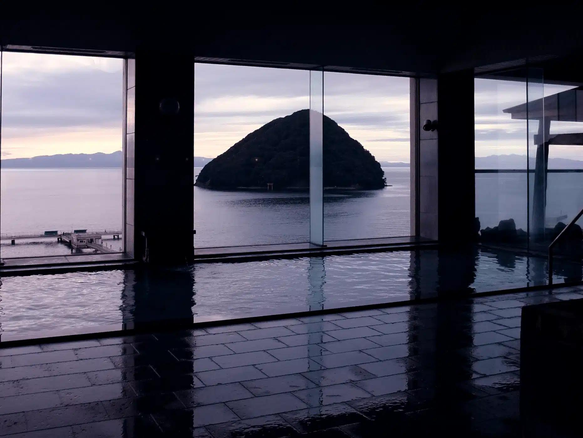 Vista desde un onsen con grandes ventanales hacia el mar al atardecer, isla rocosa en el agua y piscina interior humeante