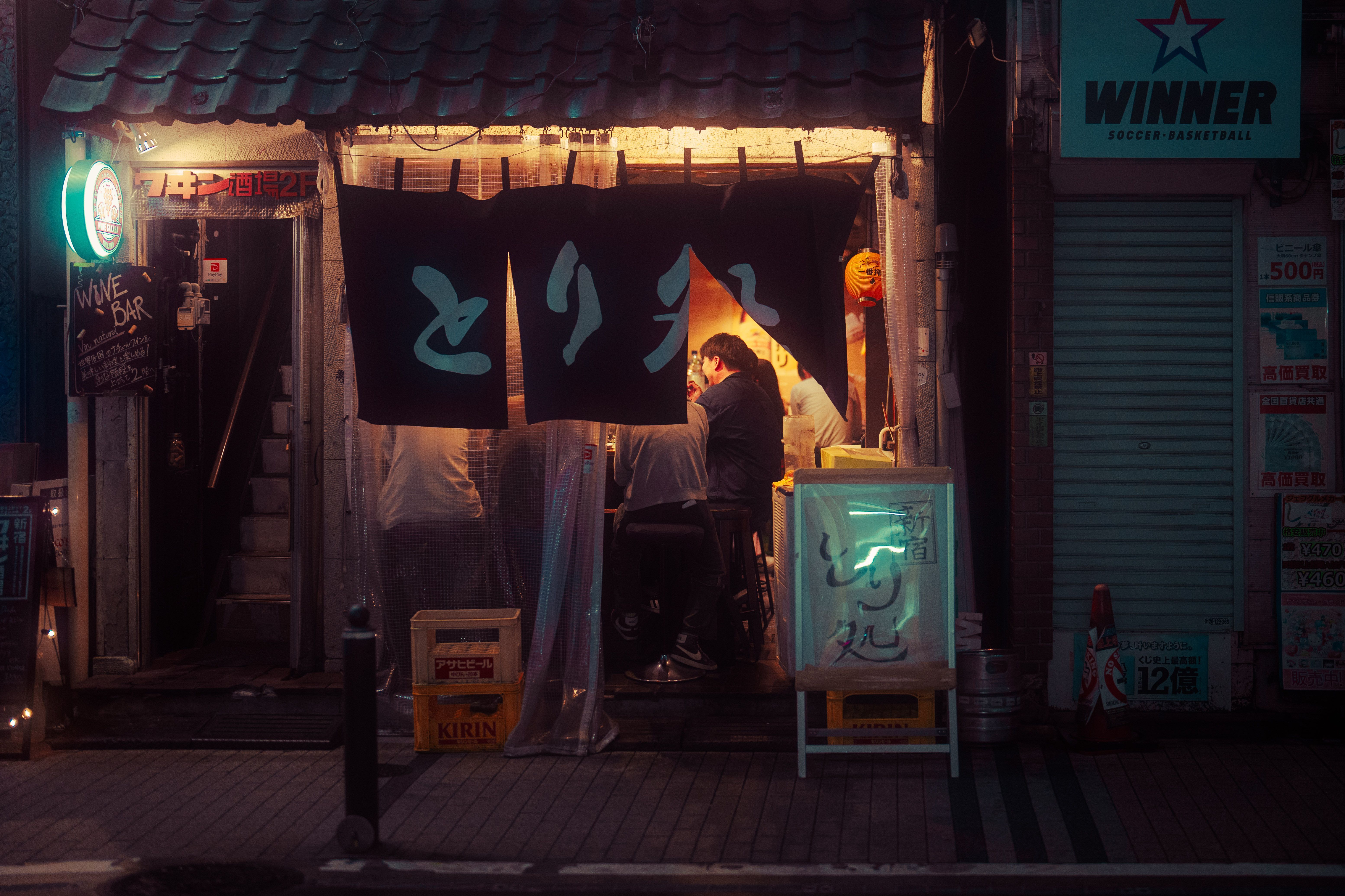 Fachada nocturna de un pequeño bar japonés con cortinas noren y personas en el interior.