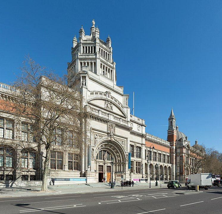 Fachada ornamentada del Museo Victoria y Albert, edificio de piedra blanca con torres y arcos victorianos, bajo un cielo azul, con árboles y peatones