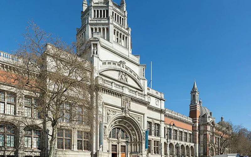 Fachada ornamentada del Museo Victoria y Albert, edificio de piedra blanca con torres y arcos victorianos, bajo un cielo azul, con árboles y peatones