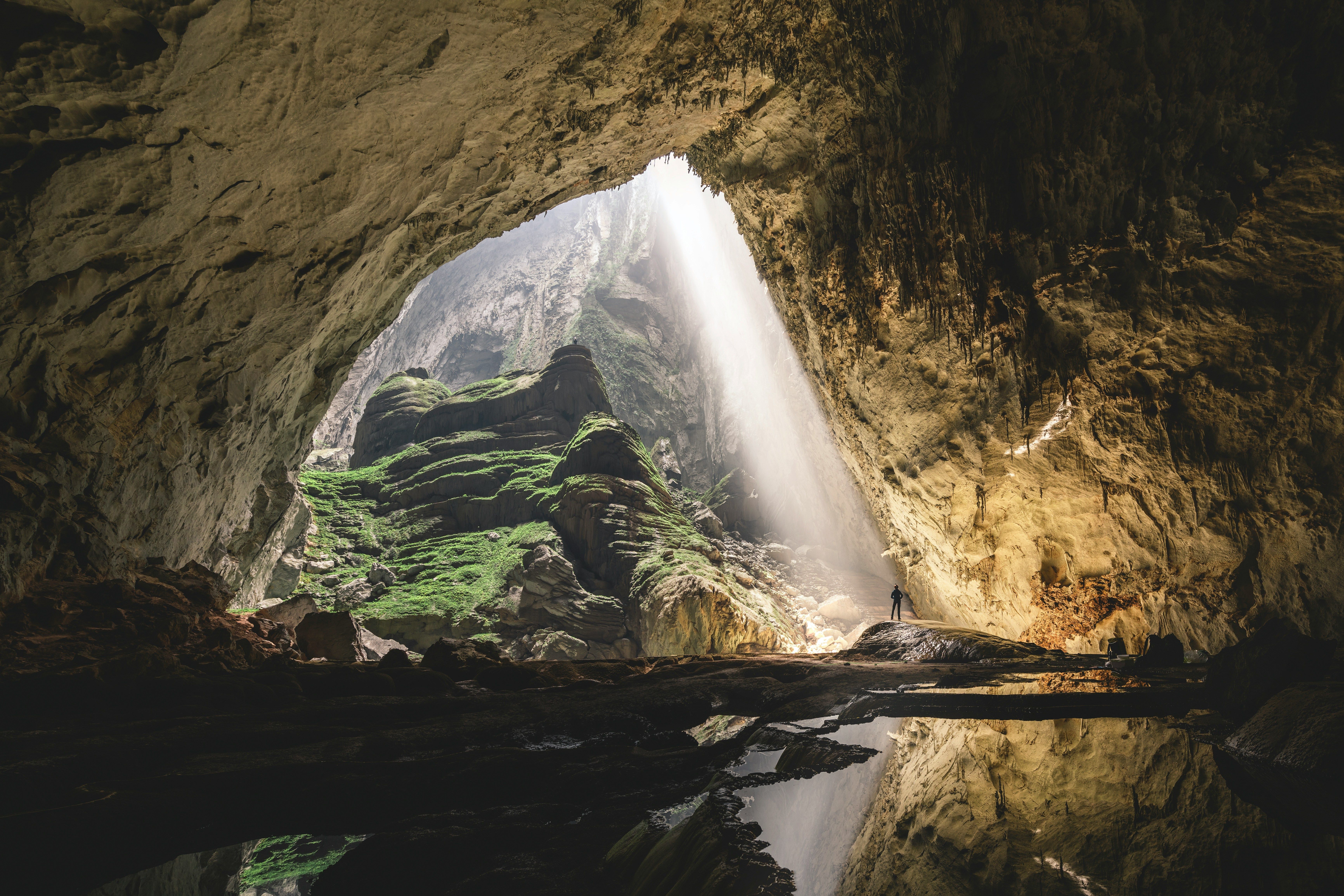 Interior de una cueva con una gran abertura superior por donde entra un potente haz de luz, iluminando el suelo y una pequeña figura humana.