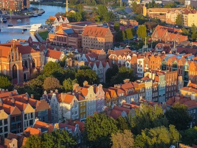 Vista panorámica de Gdansk con edificios de techos rojos y fachadas coloridas a orillas del río, barcos y vegetación verde bajo luz dorada