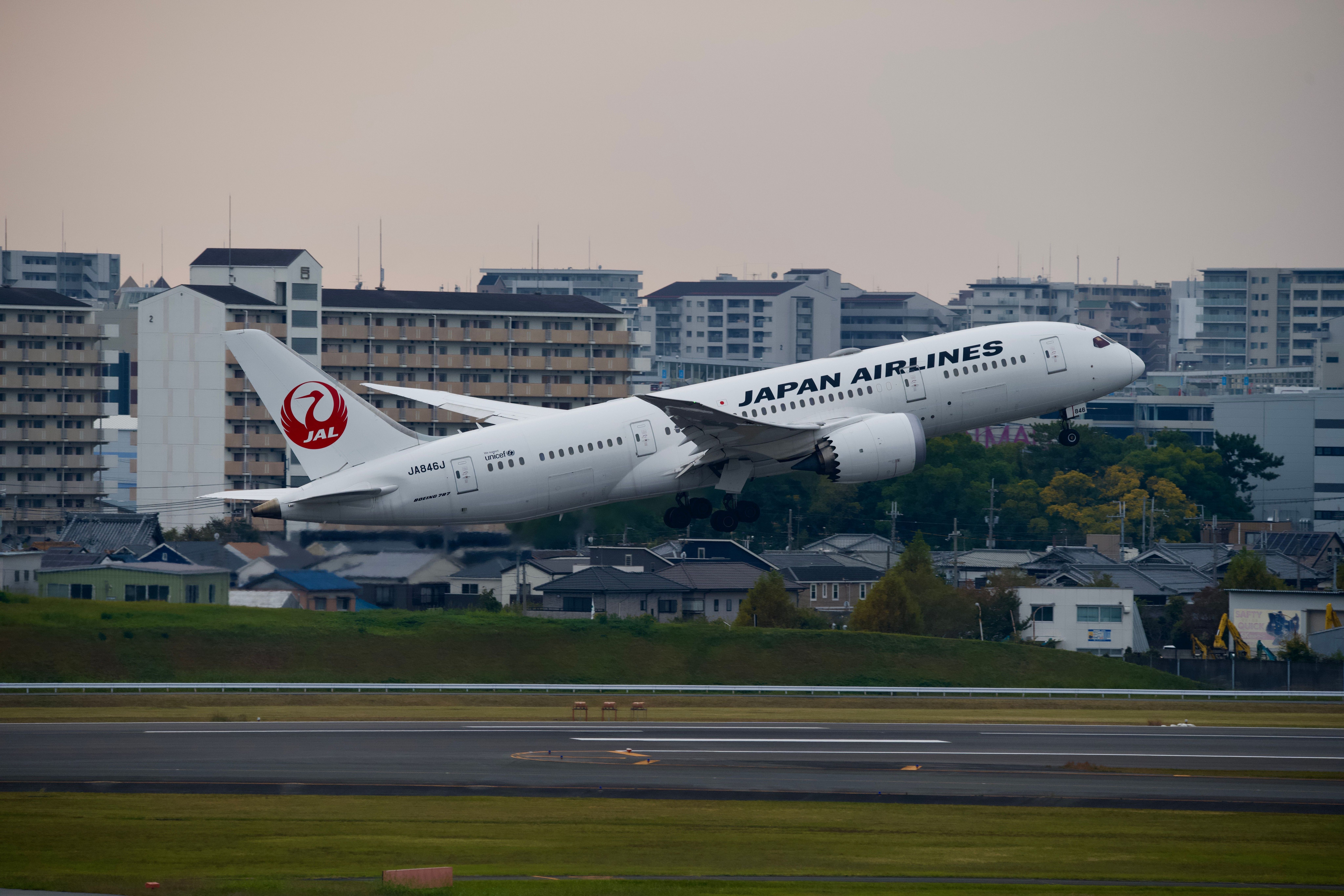 Avión blanco de Japan Airlines despegando de una pista, con edificios urbanos al fondo.