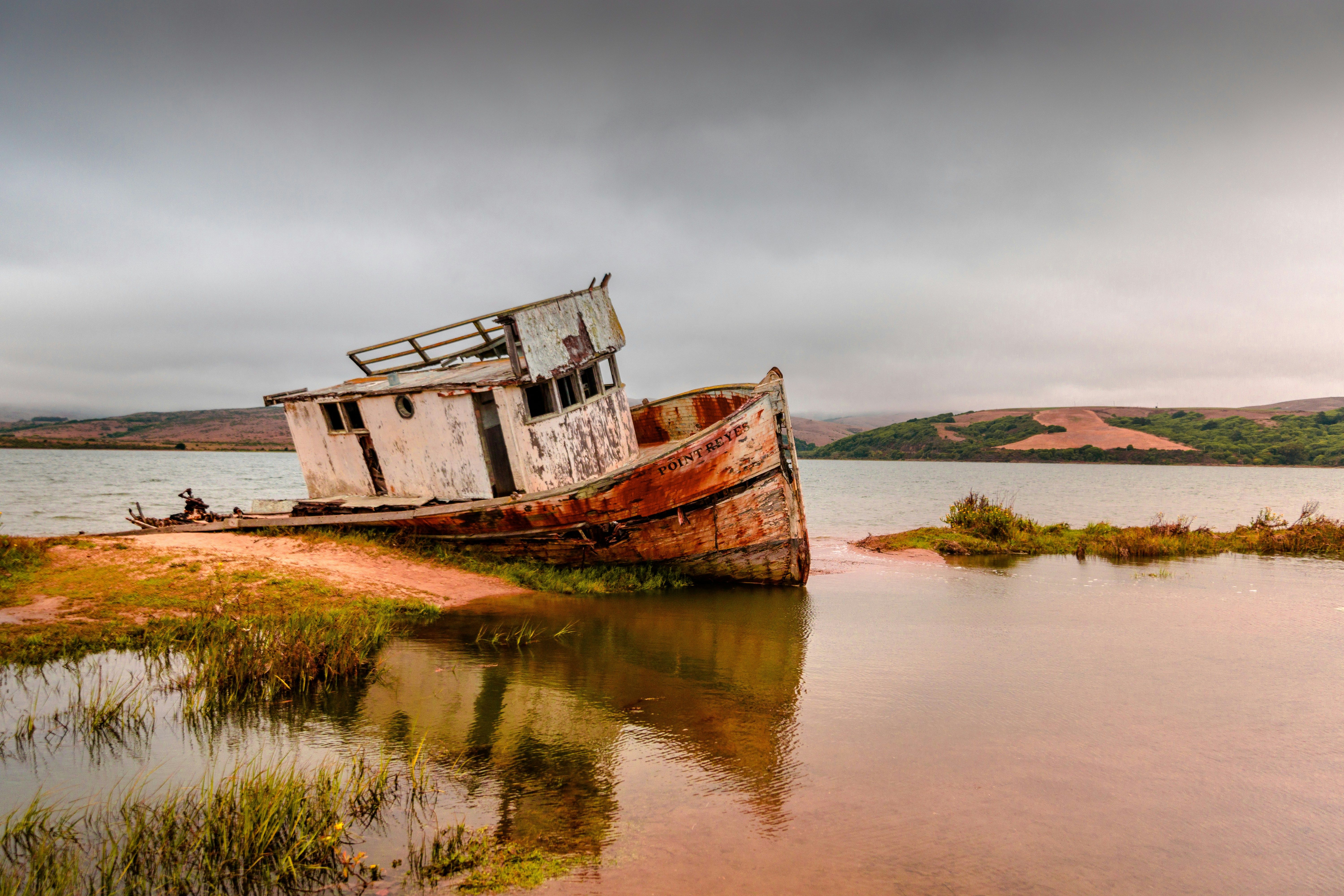 Barco de madera viejo y oxidado encallado en la orilla de un lago bajo un cielo nublado.