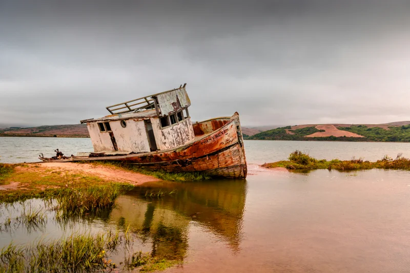 Barco de madera viejo y oxidado encallado en la orilla de un lago bajo un cielo nublado.