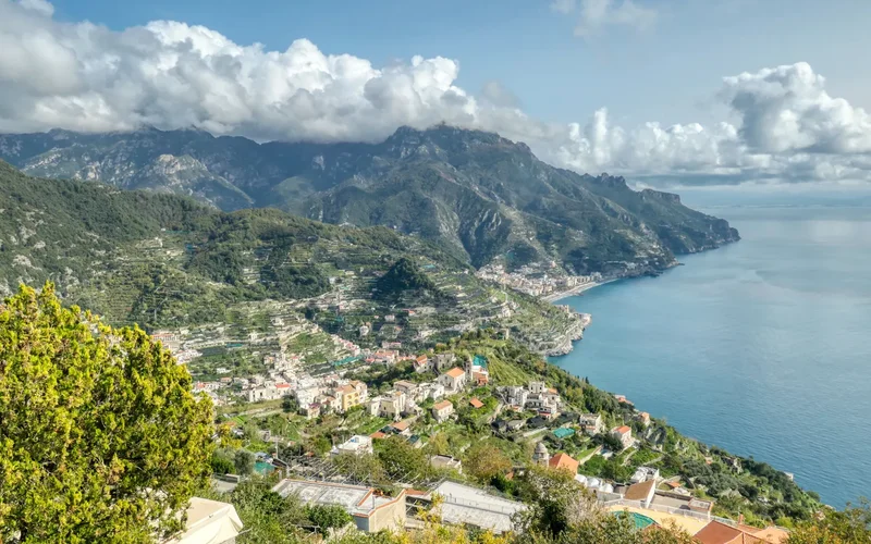 Vista panorámica de la Costa Amalfitana, con pueblos coloridos encaramados en laderas montañosas verdes, mar azul y nubes blancas en el cielo