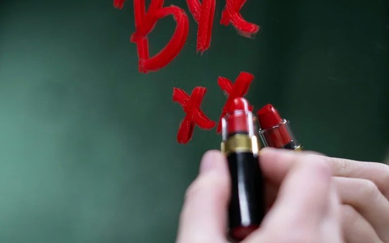 Manos sosteniendo lápices labiales dorados escribiendo "BYE XX" en rojo sobre un espejo verde