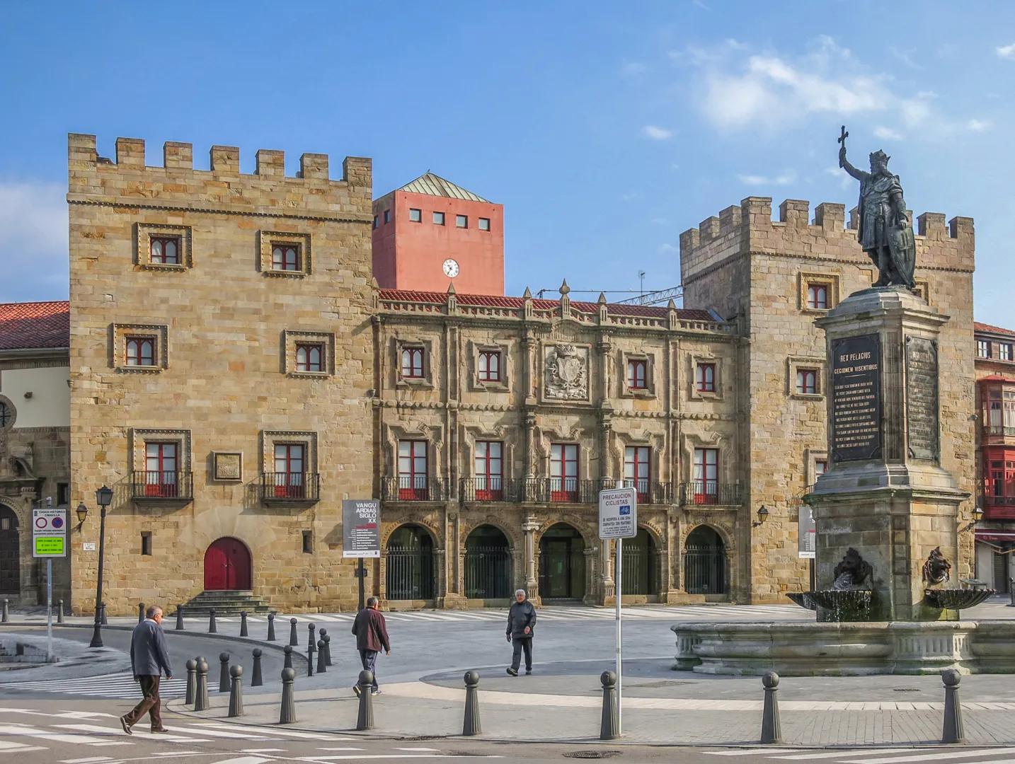 Vista de la plaza del Ayuntamiento de Gijón con su edificio histórico de piedra, estatua de Pelayo y fuente bajo cielo azul