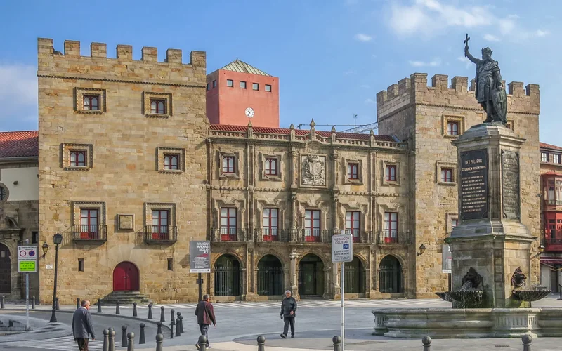 Vista de la plaza del Ayuntamiento de Gijón con su edificio histórico de piedra, estatua de Pelayo y fuente bajo cielo azul